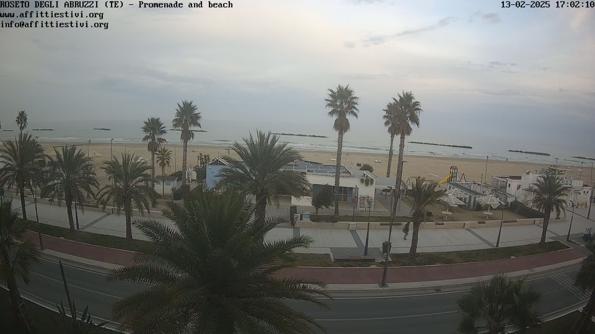 A mostly cloudy day shows a sandy beach with calm waves, palm trees lining a paved promenade and road next to it.
