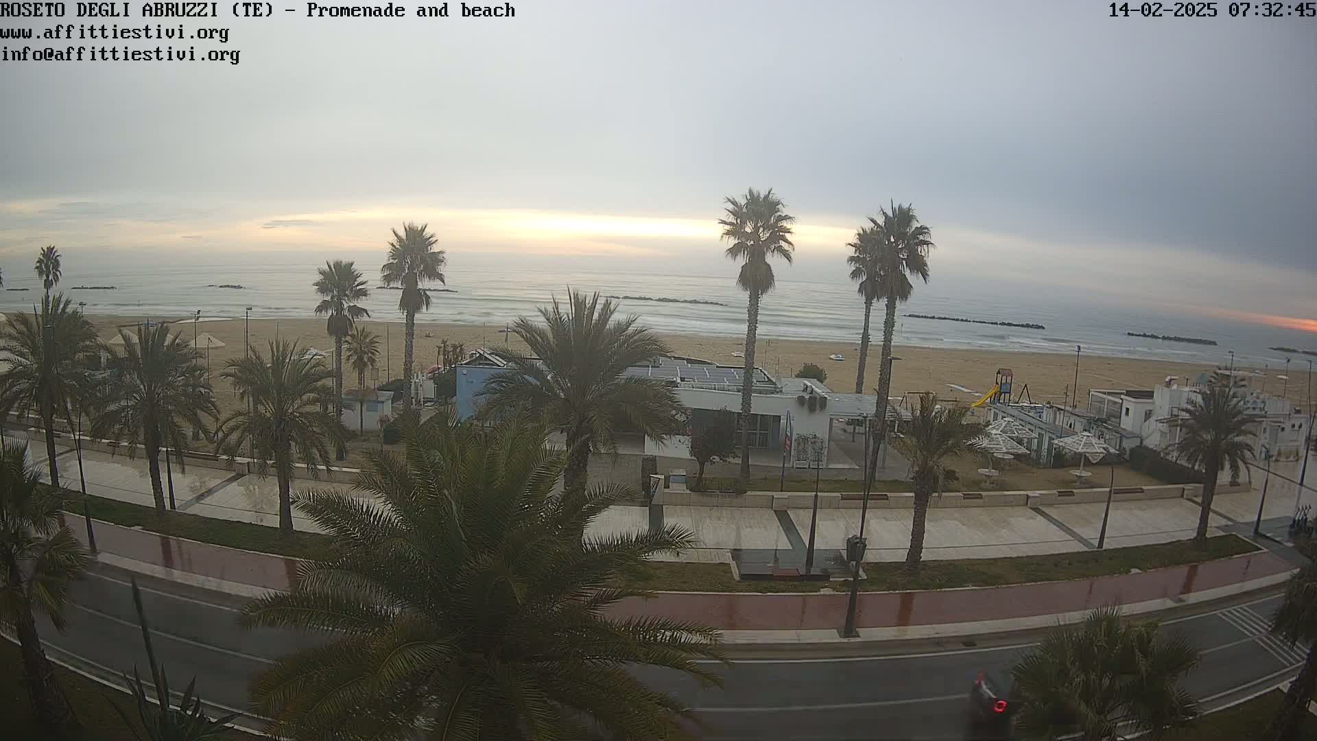 A mostly cloudy sunrise illuminates a beach with palm trees lining a paved promenade and road.