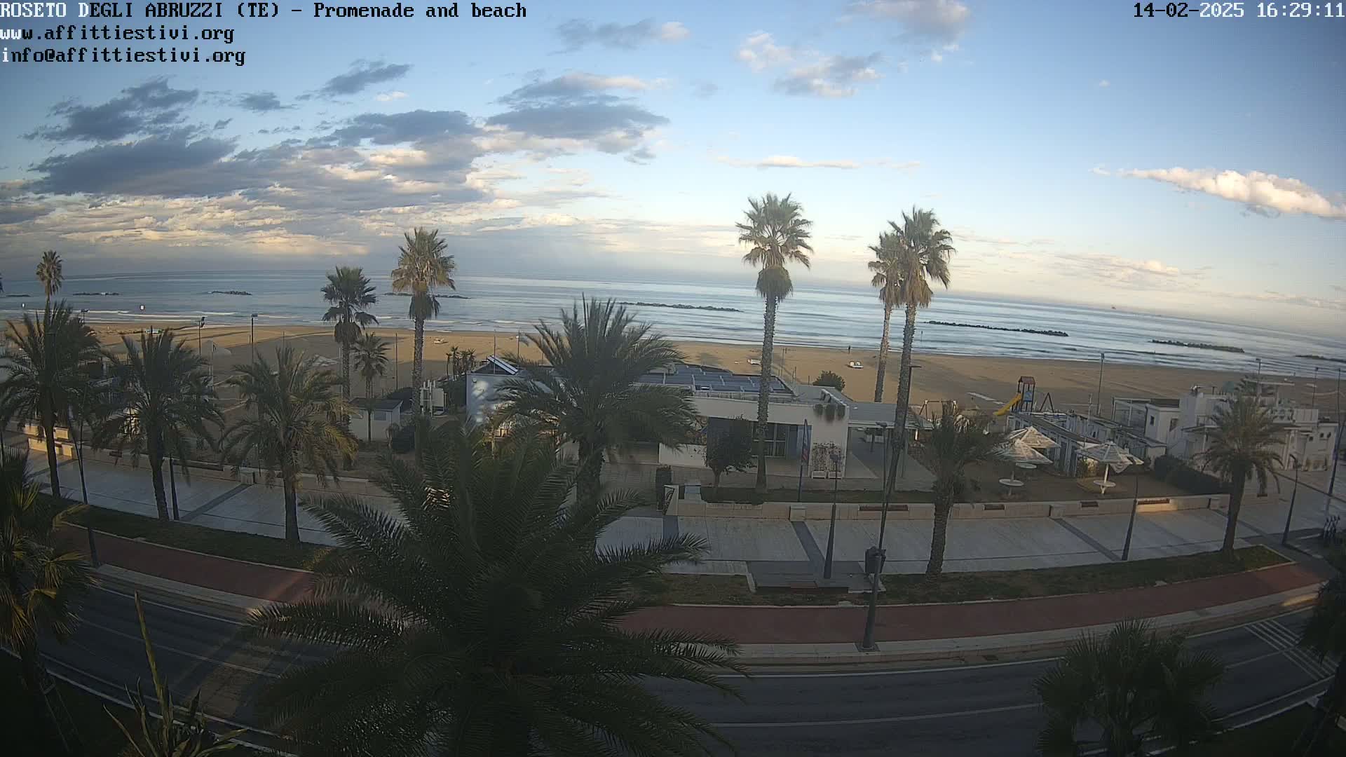 A sandy beach with calm waves and palm trees lining a promenade next to a road under a partly cloudy sky.