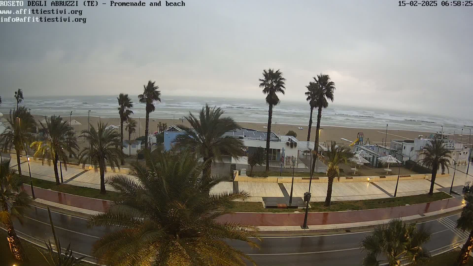 A rain-soaked promenade and beach scene features palm trees lining a roadway overlooking a stormy sea with choppy waves.