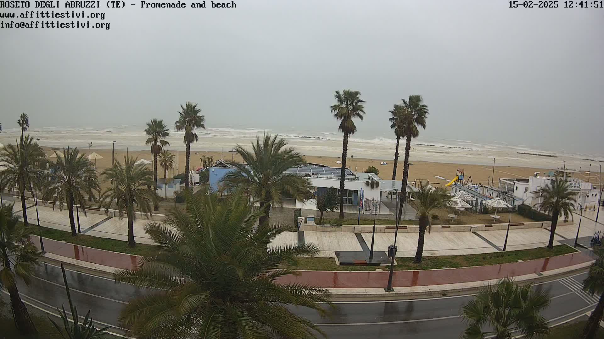 A wet, overcast day reveals a beach with palm trees lining a promenade, overlooking a turbulent sea.
