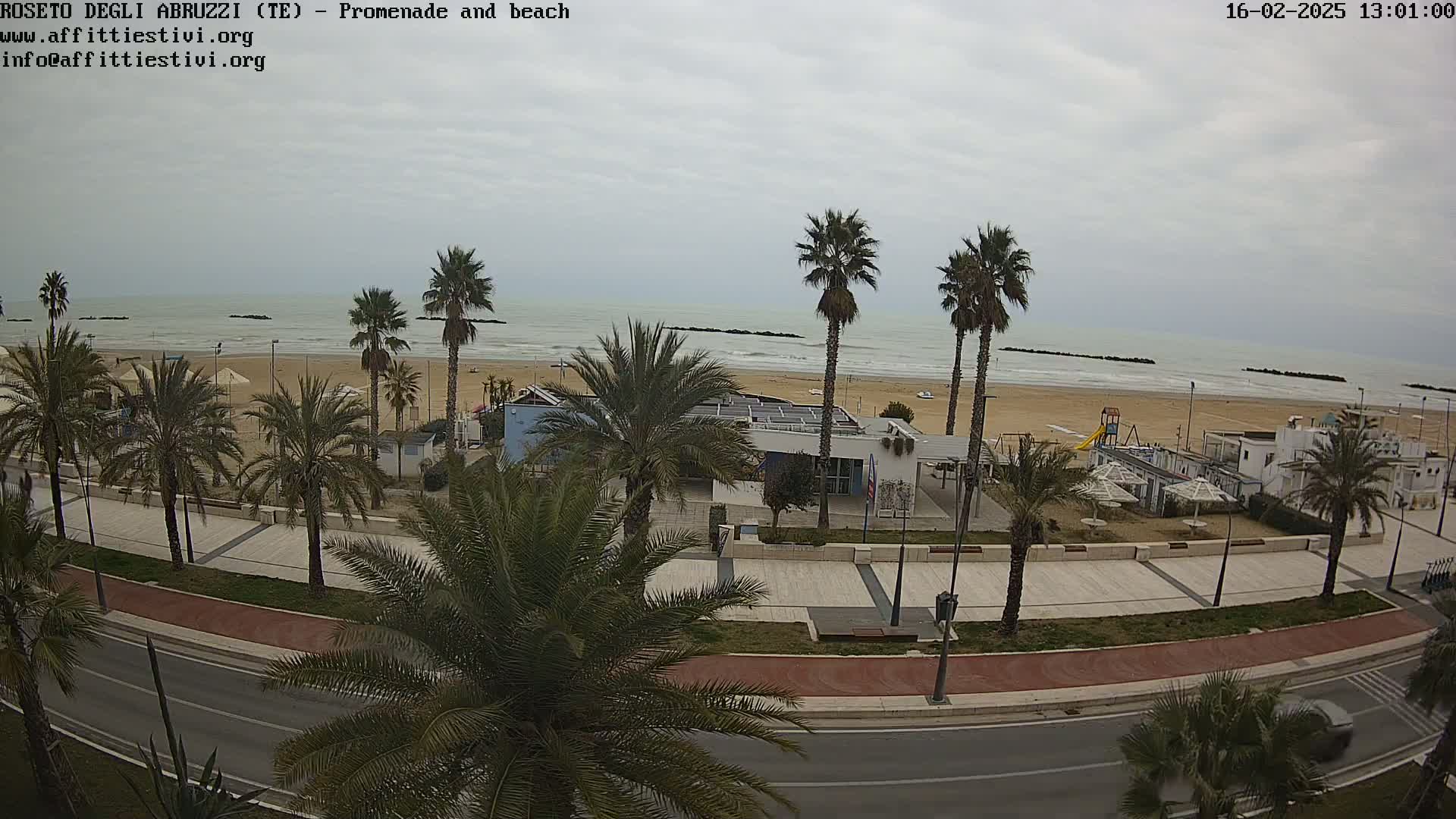 A beach with calm waves under an overcast sky is seen from above, showing a promenade lined with palm trees and beachside buildings.