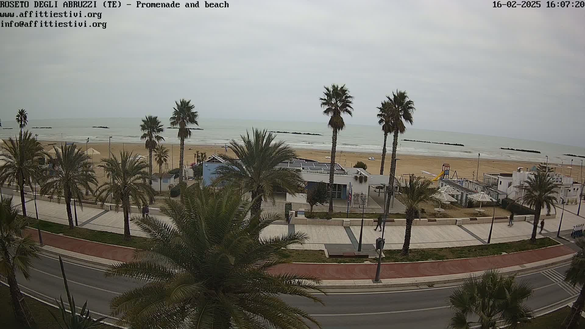 A beach with calm, gray water is seen from above, with a promenade lined with palm trees, a road, and some buildings.