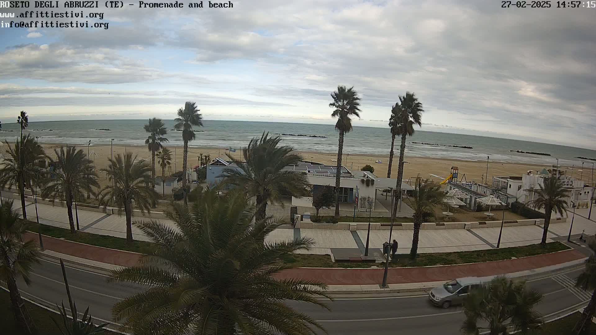 A seaside promenade with palm trees lines a paved walkway next to a sandy beach and calm ocean under an overcast sky.