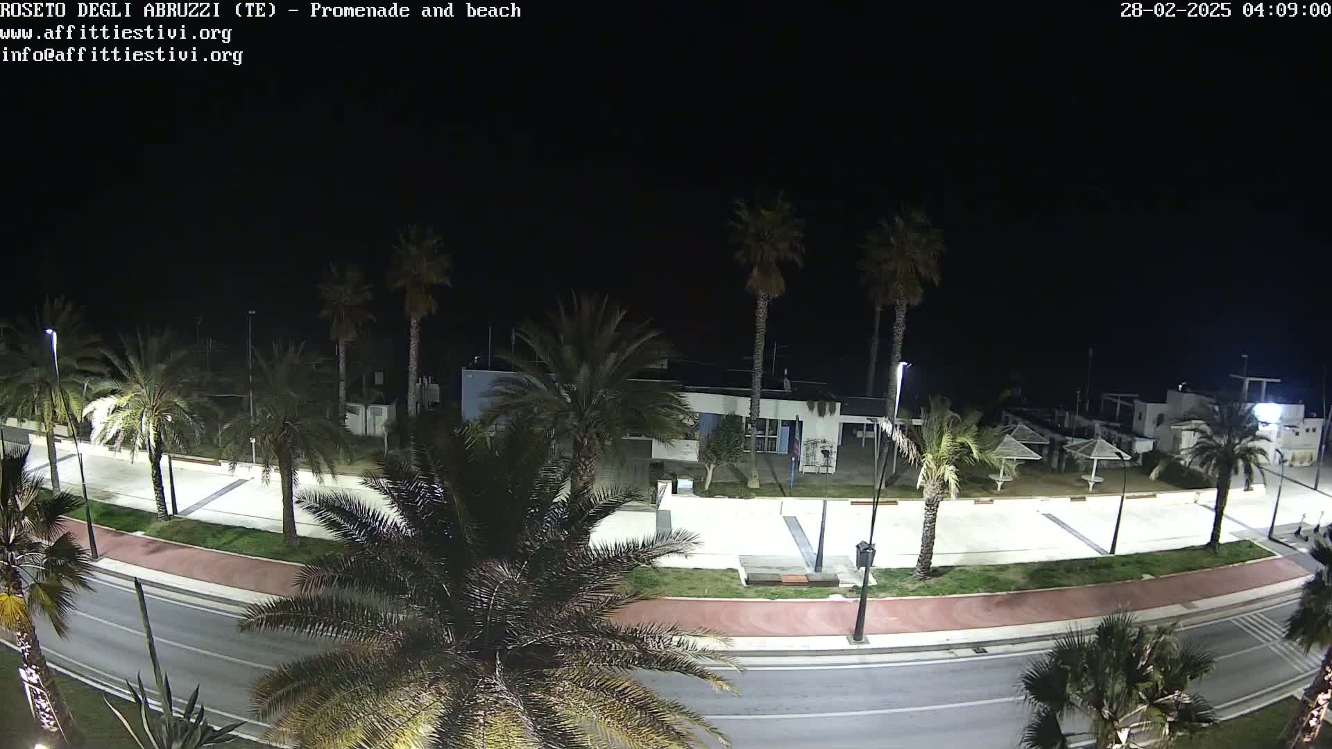 A nighttime view of a paved promenade lined with palm trees, leading to a beach area with a few closed beach umbrellas and a building under a dark sky.