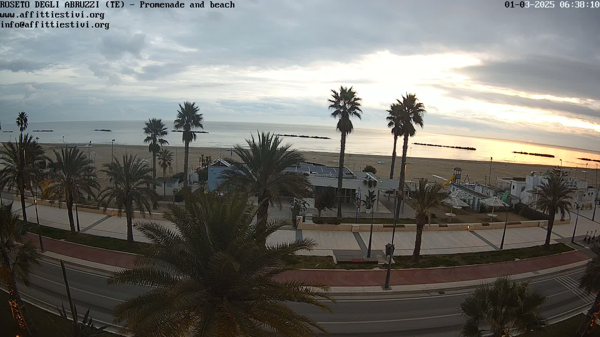 A seaside promenade lined with palm trees overlooks a sandy beach and calm ocean under a partly cloudy sunrise sky.