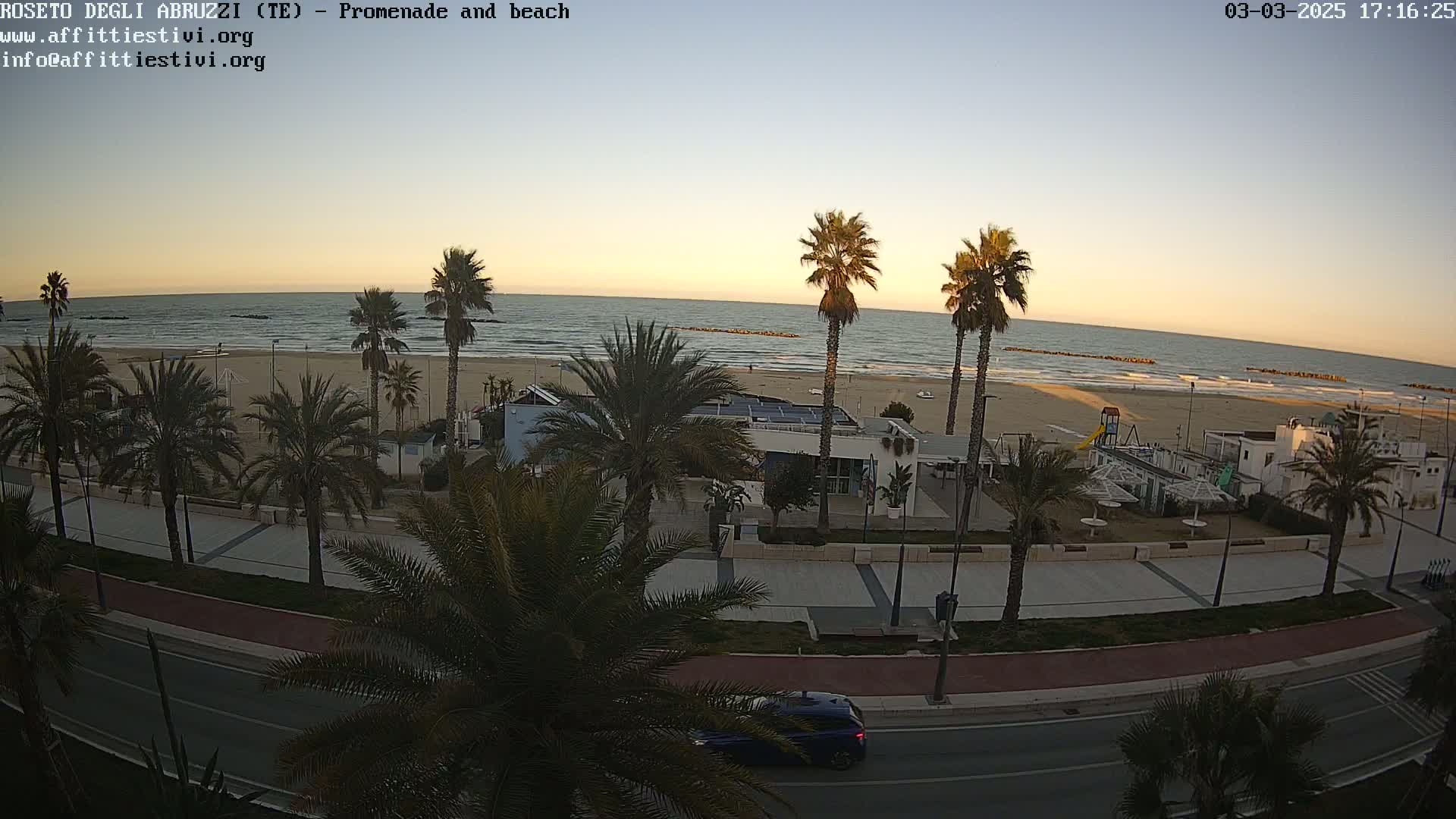 A beach with calm waves at sunset is viewed from above, showing a promenade lined with palm trees, buildings, and a road with a single car.