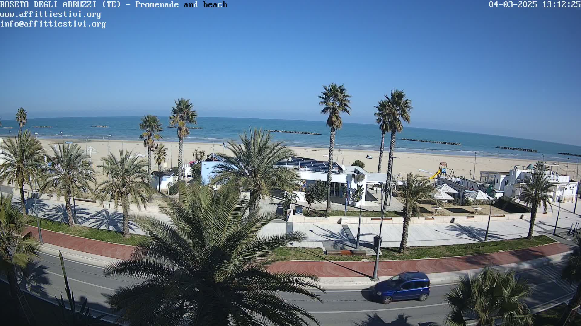 A sunny day overlooking a beach with palm trees lining a promenade and a road, with a single car visible.