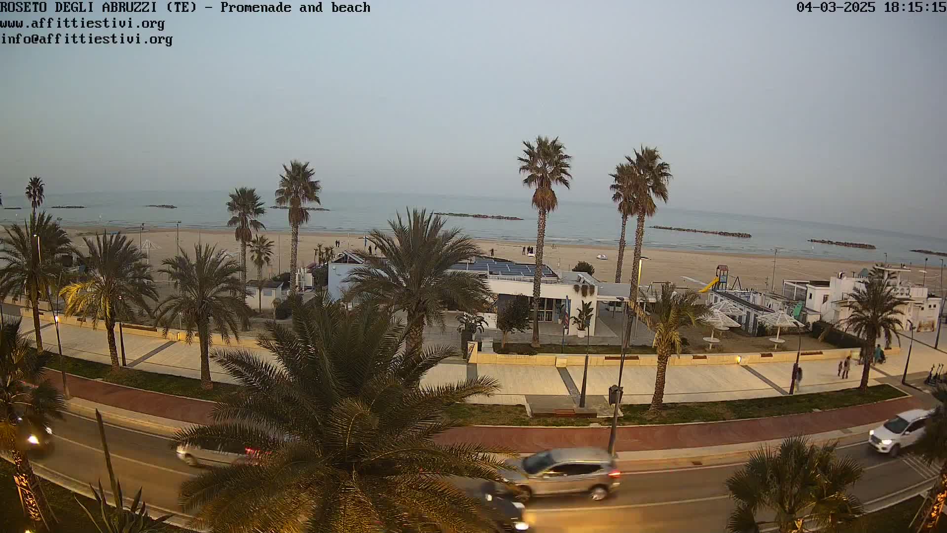 A seaside promenade at dusk, lined with palm trees, overlooks a sandy beach and calm ocean under a pale sky.