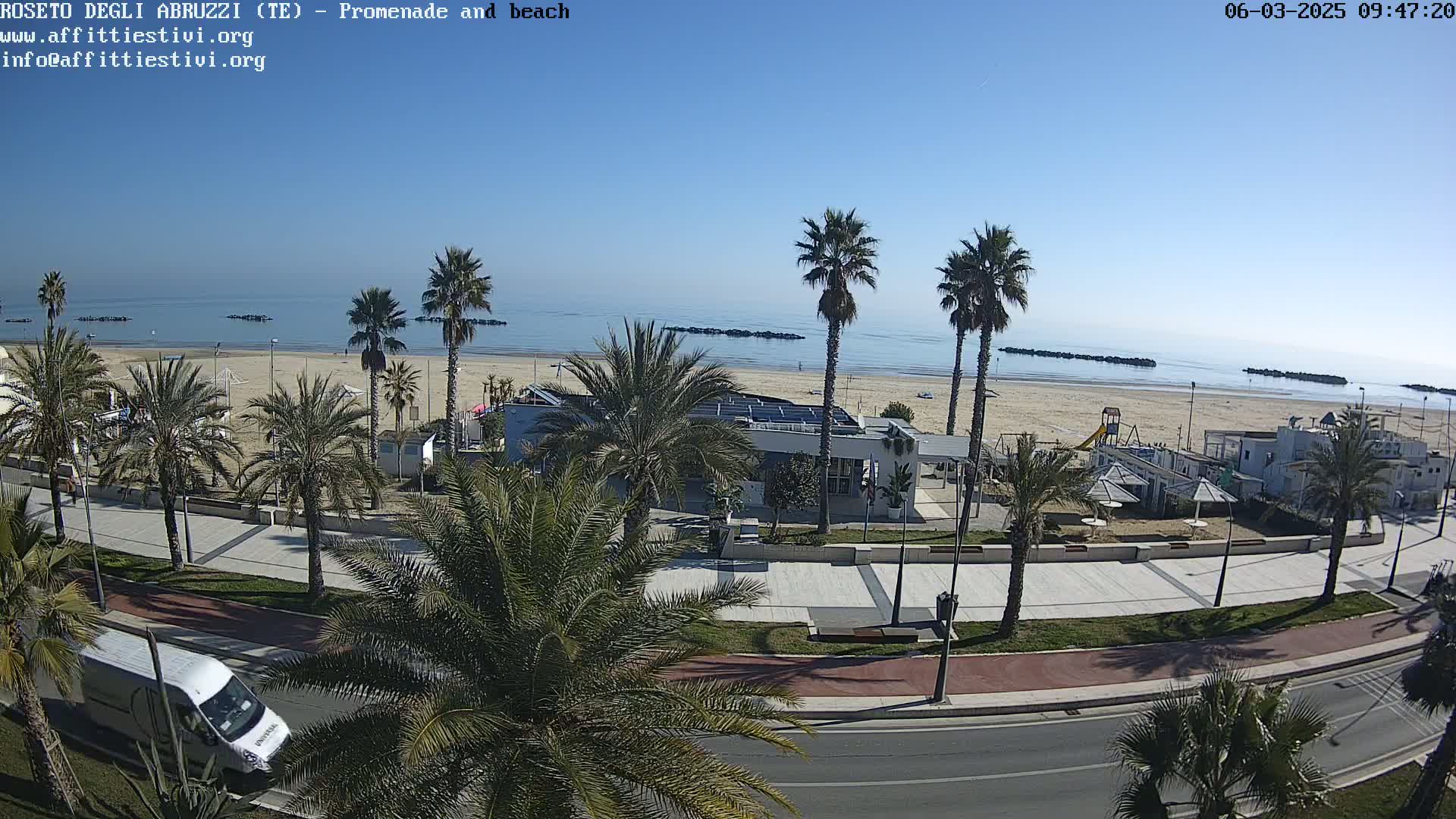 A beach with calm water under a clear blue sky is visible from a high vantage point, showing a promenade lined with palm trees and beachside buildings.
