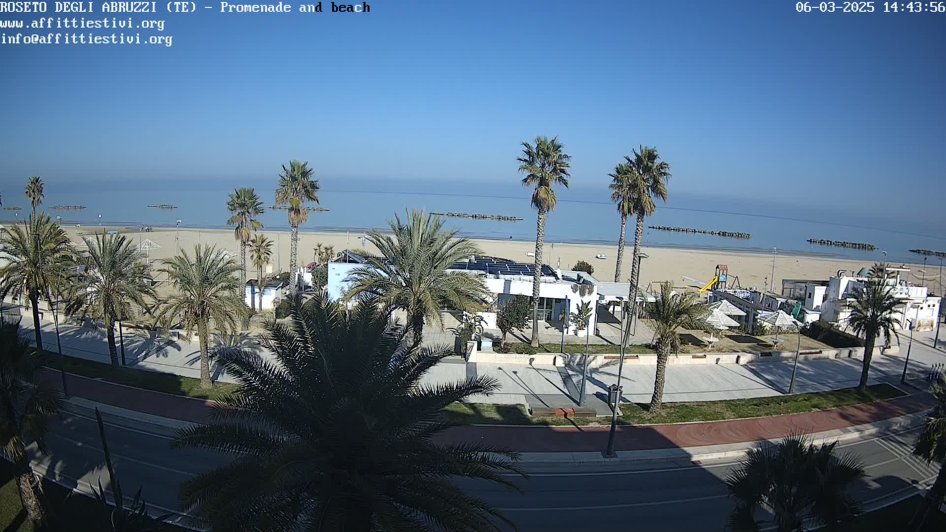A sunny day overlooking a beach with palm trees lining a paved promenade and road, next to buildings and beach equipment.