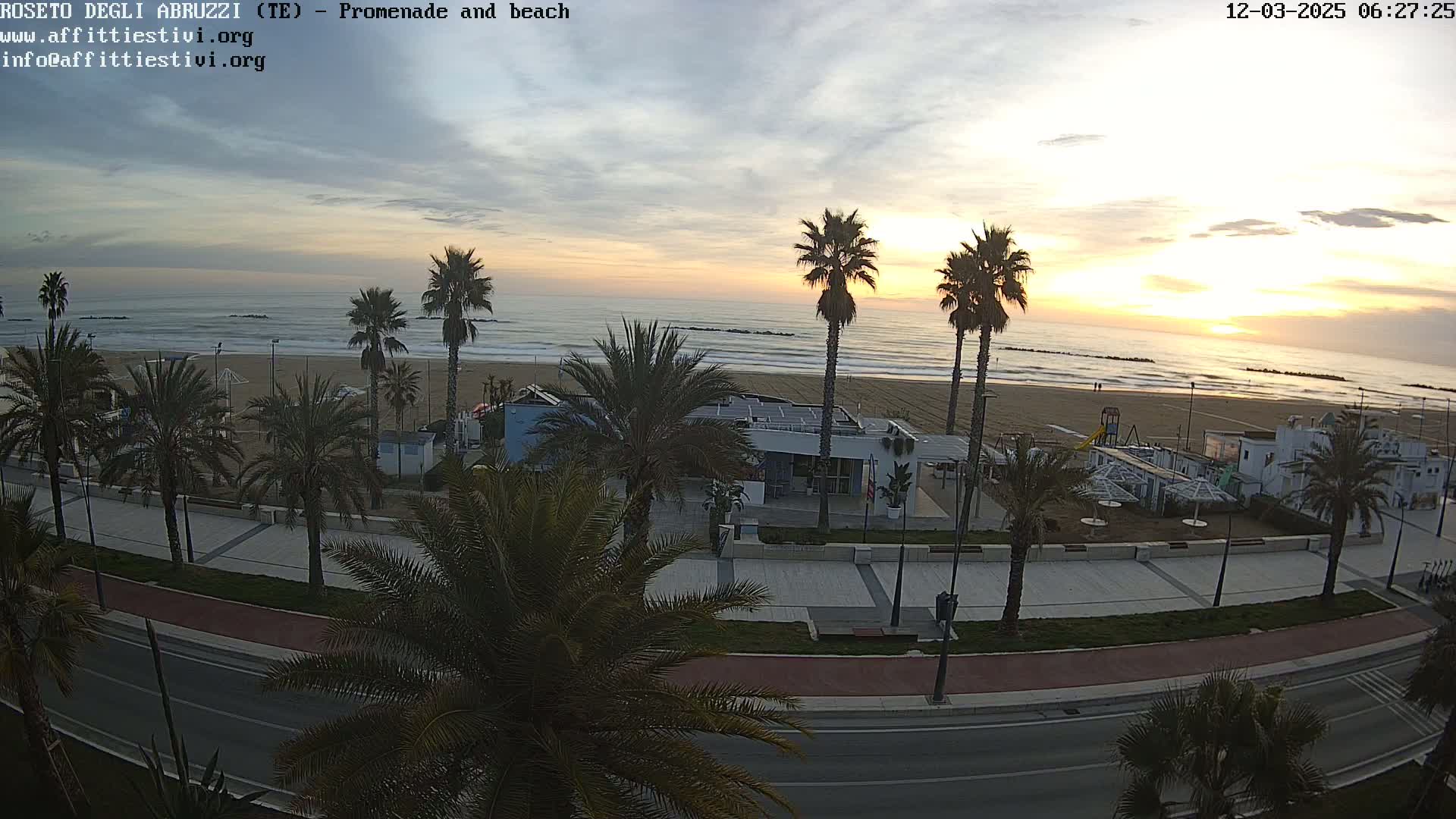 A sunrise over a beach with palm trees lining a promenade and road, under partly cloudy skies.