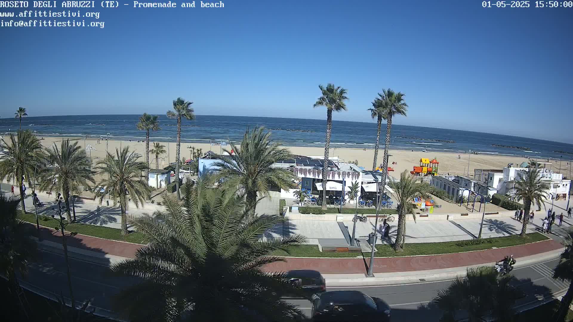 A sunny beach scene shows a promenade lined with palm trees, a beachfront restaurant, and a playground, with cars driving on a road nearby.