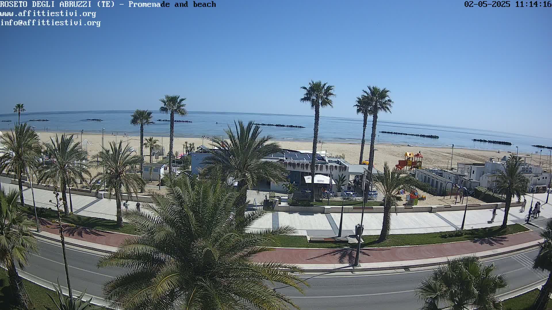 A sunny beach with palm trees lining a paved promenade and a road,  with a small playground and beachside buildings visible.