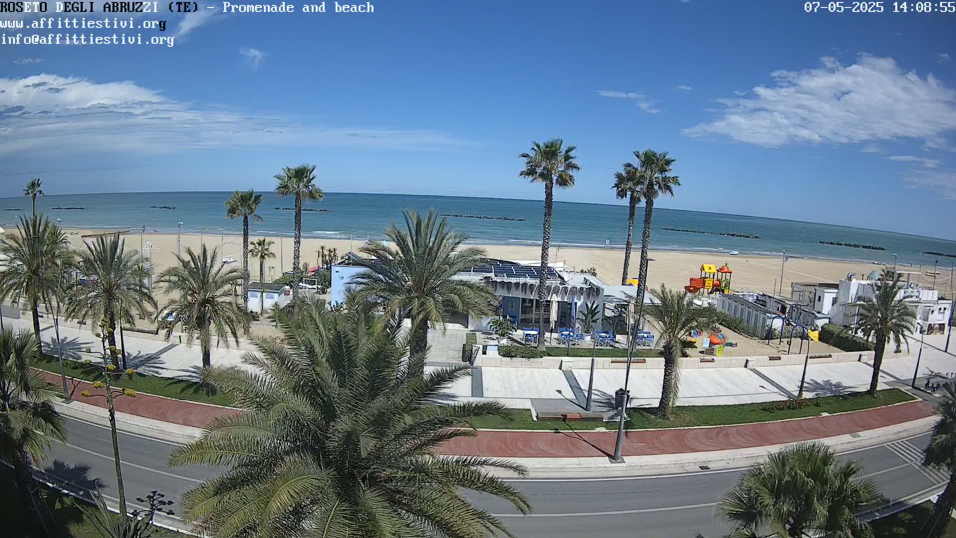 A beachside promenade with palm trees, a beach with calm blue water under a mostly sunny sky, and a playground is visible from an elevated viewpoint.