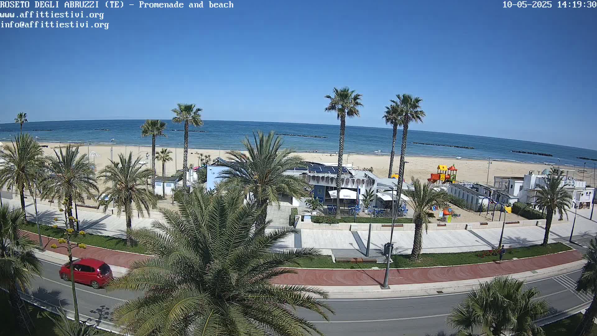 A sunny day at a beachside promenade shows palm trees lining a paved path next to a sandy beach with a calm ocean, and a red car driving on the road.