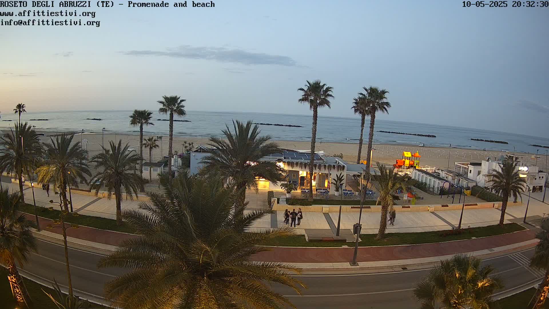 A seaside promenade at dusk, lined with palm trees, overlooks a sandy beach and calm ocean.