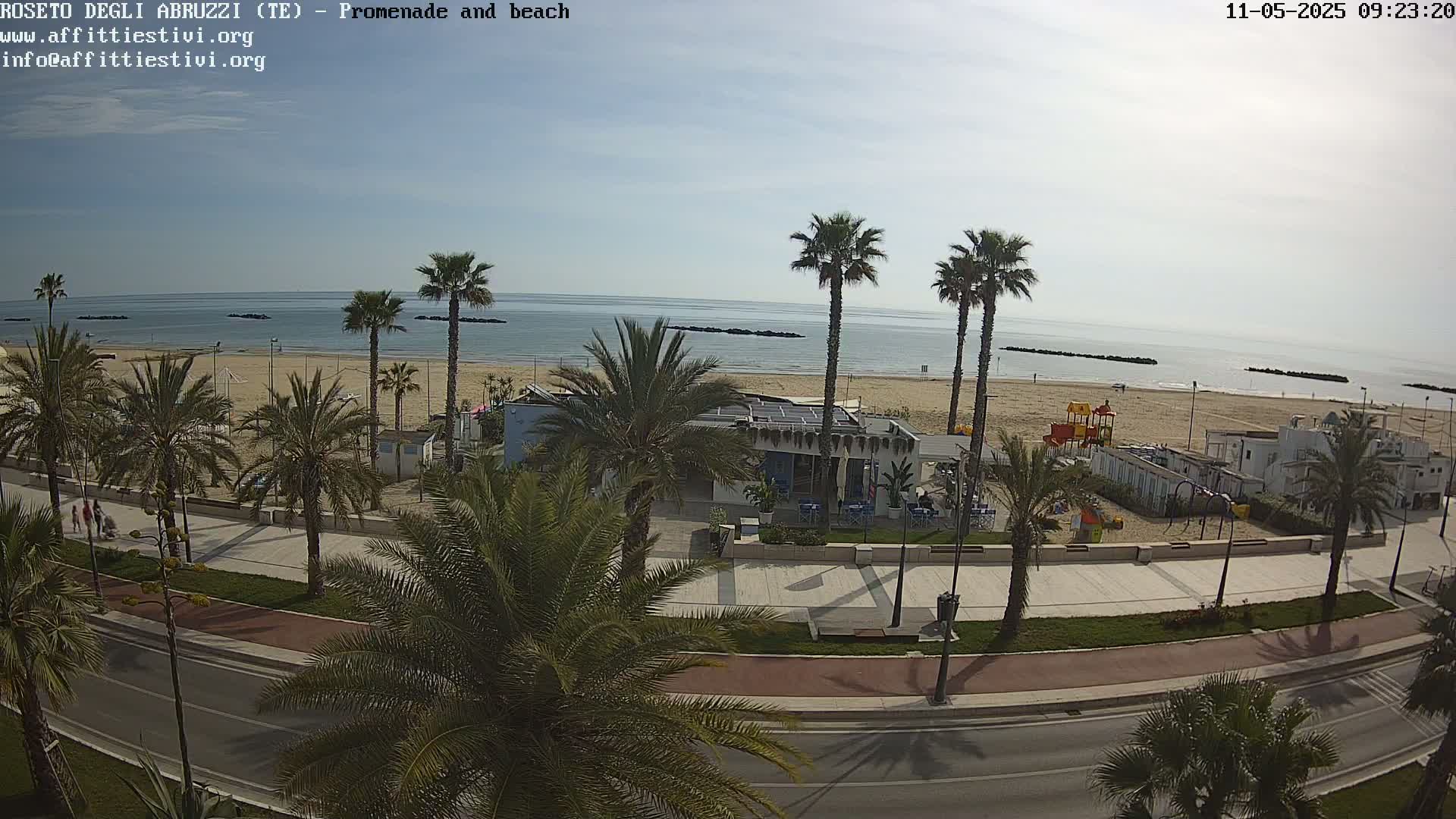 A sunny day overlooking a beach promenade lined with palm trees, a beachfront building with outdoor seating, and a playground, with a calm sea in the background.