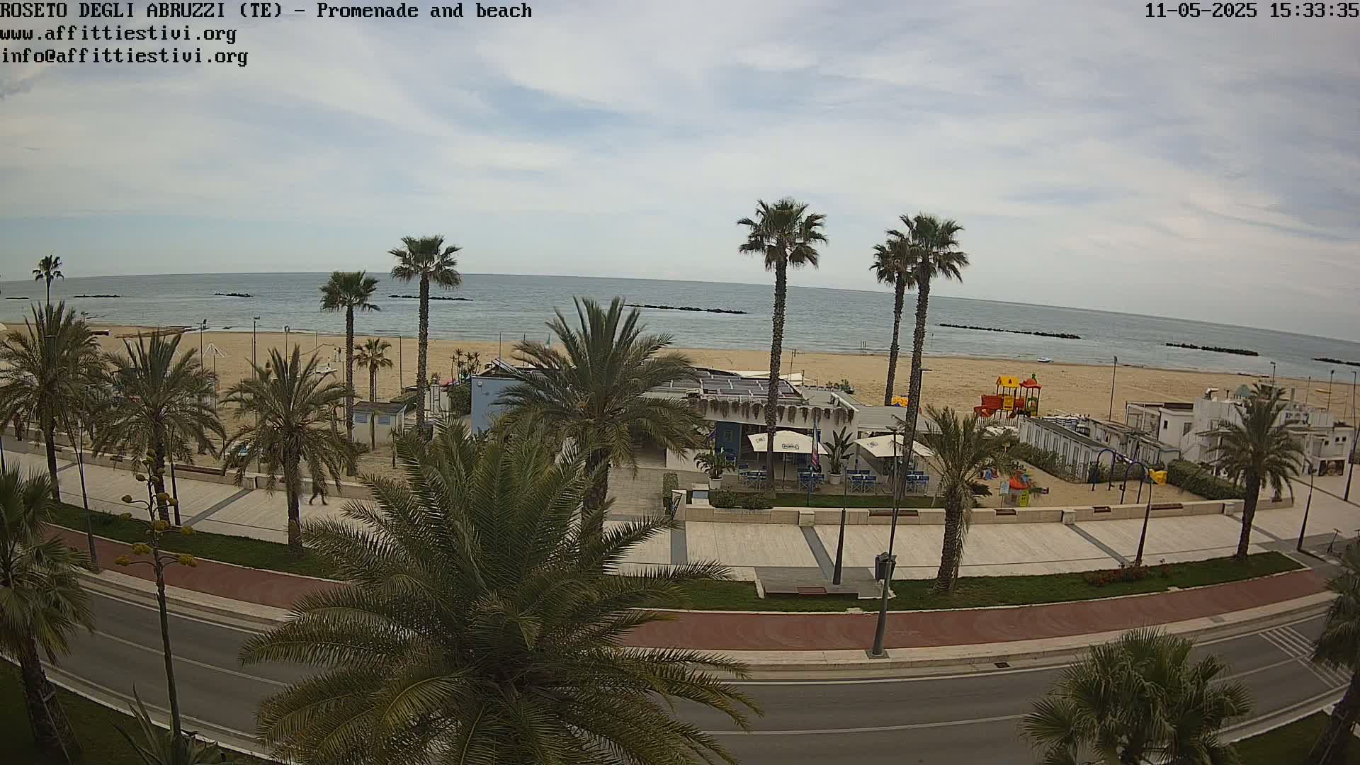 A beach with calm water under a cloudy sky is viewed from above, showing a promenade lined with palm trees, a beachfront restaurant, and a children's playground.