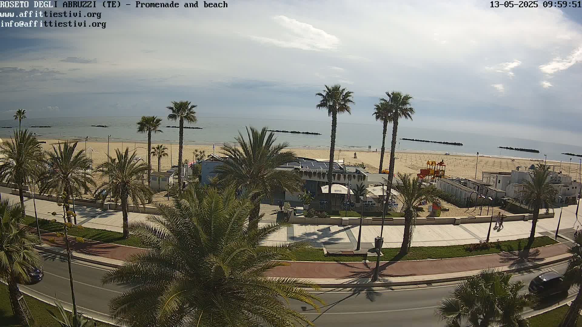 A partly cloudy day shows a beach with palm trees lining a promenade and road next to a calm ocean.