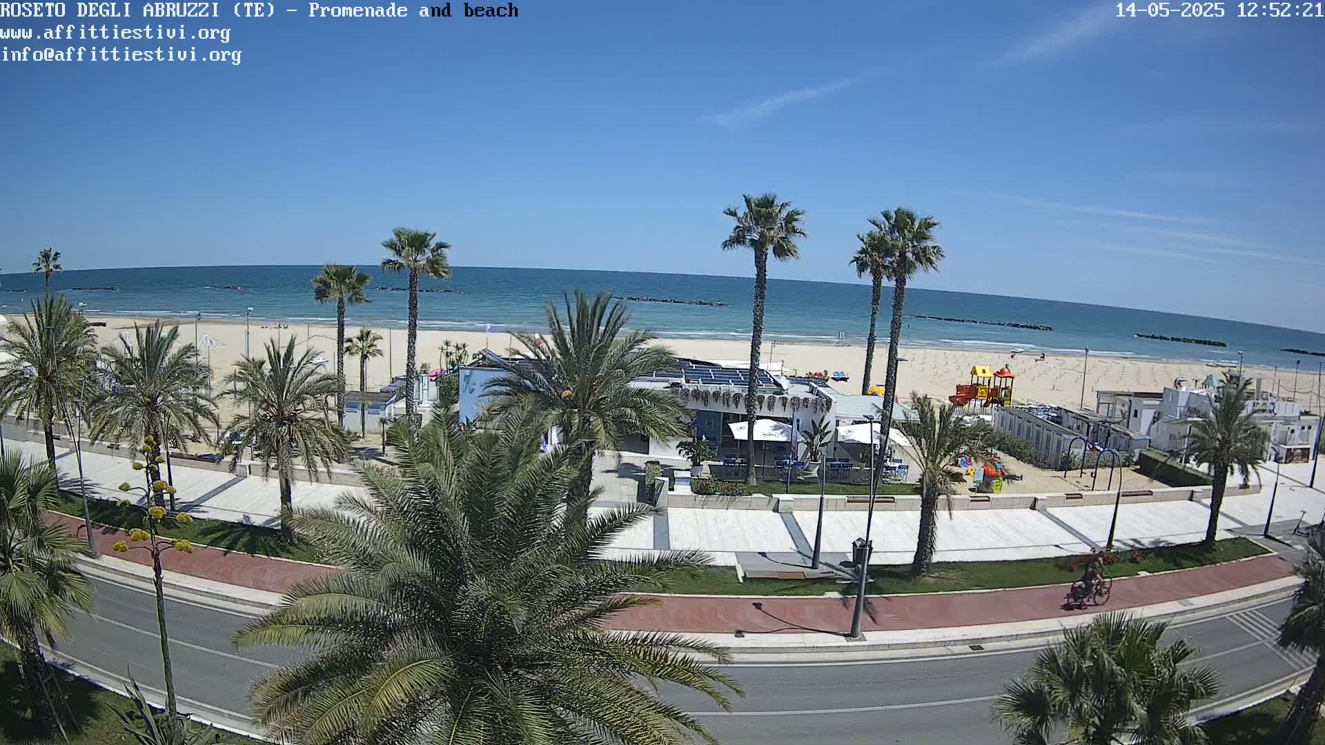 A beach scene on a sunny day shows a sandy beach with people and beach umbrellas, palm trees lining a paved promenade and road next to the beach, a beachside cafe, and a playground.