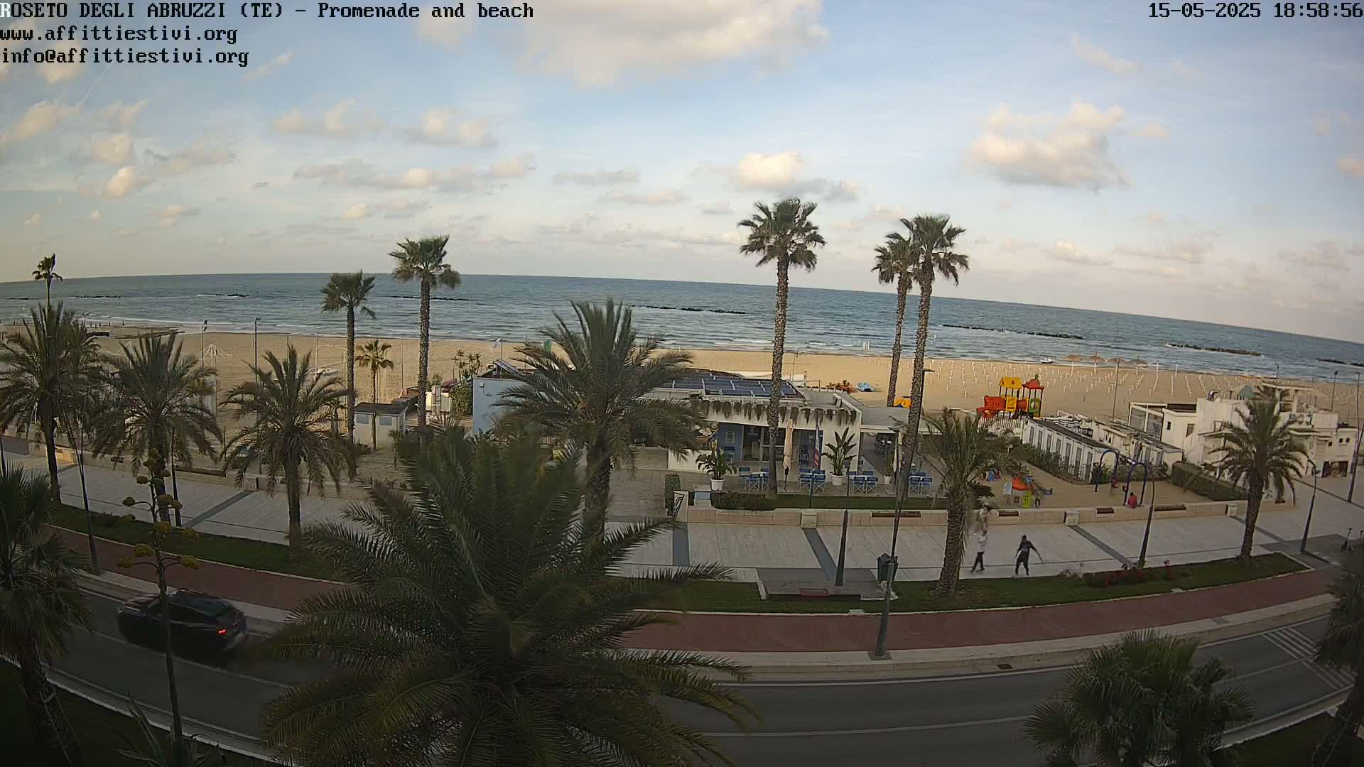 A beach with calm ocean waves, palm trees lining a promenade, and a playground is visible under a partly cloudy sky.