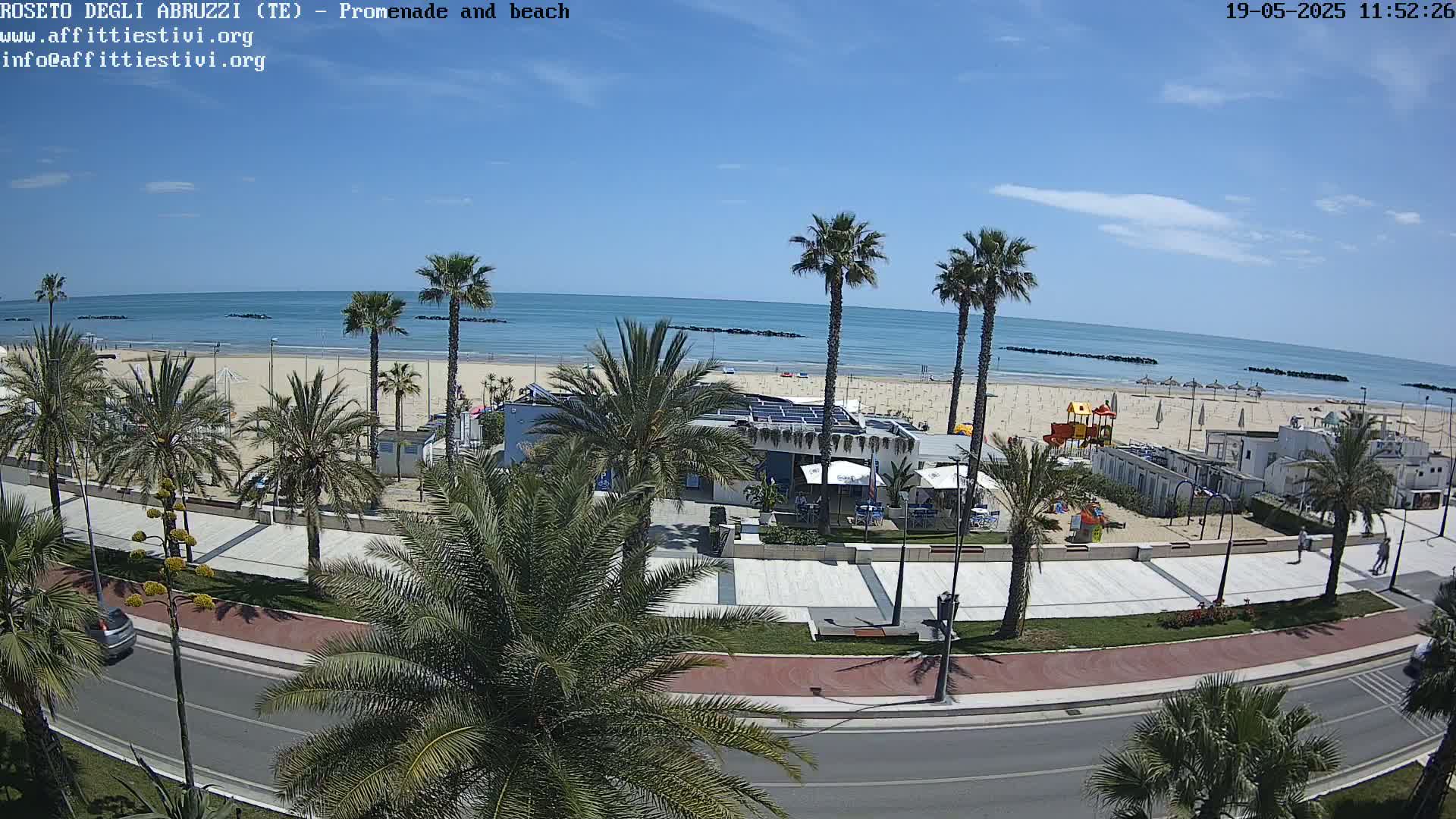 A beach with calm, blue water and a promenade lined with palm trees is visible on a sunny day.