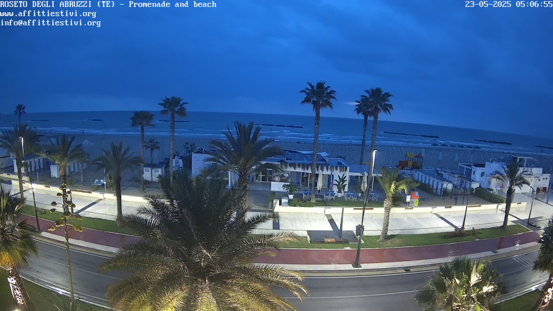 A seaside promenade at twilight shows a calm ocean, sandy beach, palm trees lining a paved walkway, and beachside buildings.