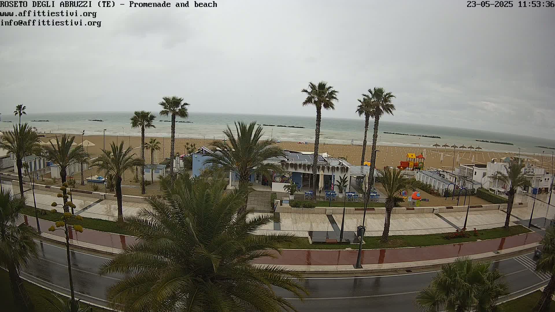 A wet, overcast day at a beachside promenade shows palm trees lining a road next to a sandy beach with a few beach umbrellas and a playground.