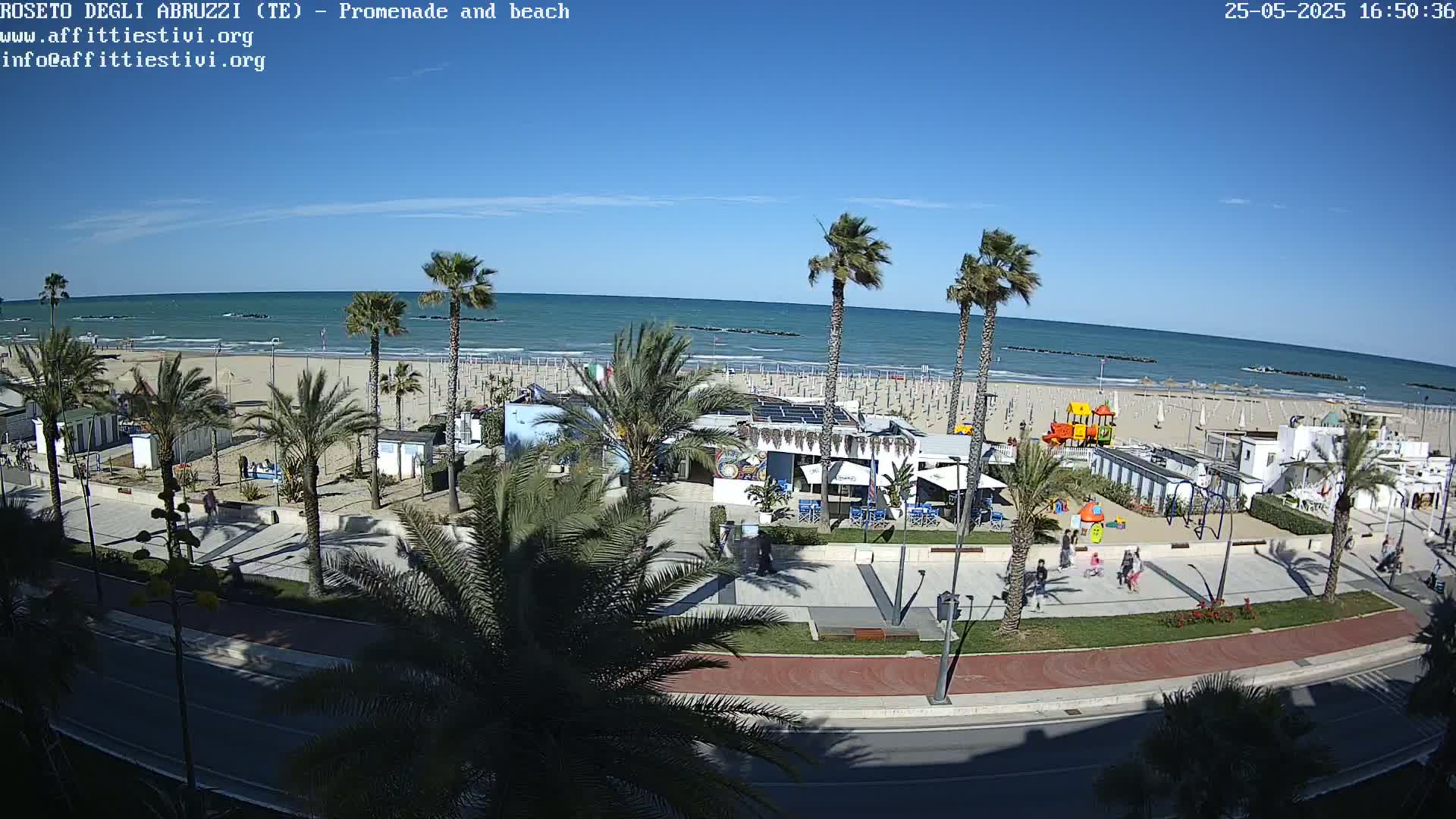 A sunny beach scene shows a promenade lined with palm trees, beachfront buildings, and a playground, overlooking a calm ocean under a clear blue sky.