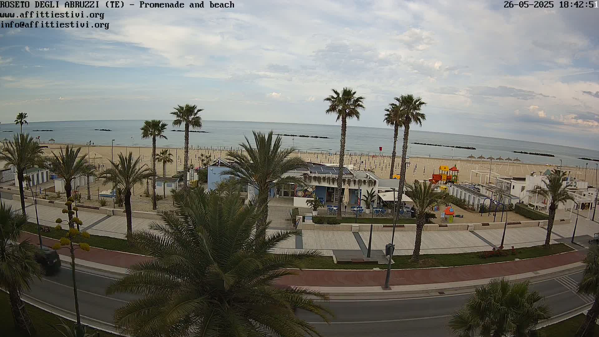 A beach with palm trees, a promenade, and a seaside restaurant is visible under a partly cloudy sky.