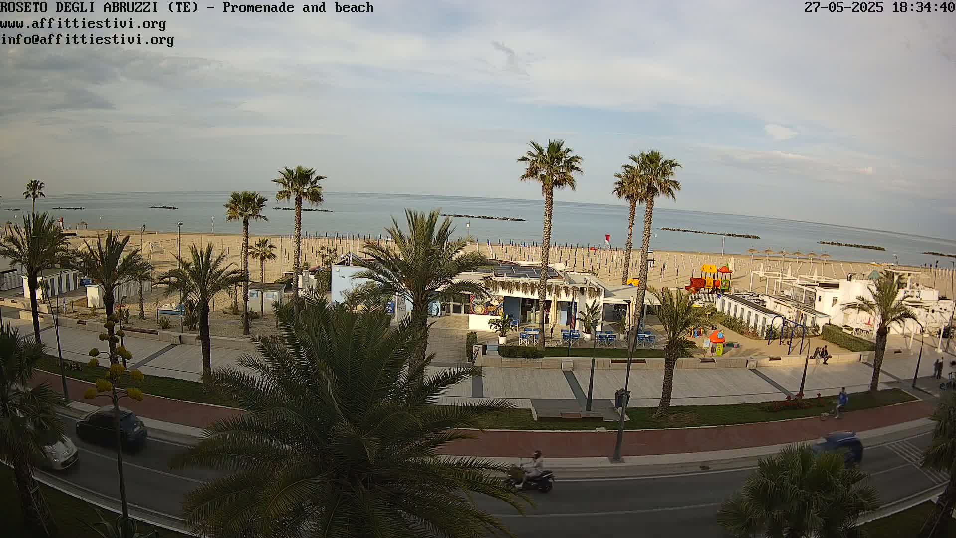 A beach with calm water and palm trees lining a promenade with cars and a person on a scooter, under a mostly clear sky.