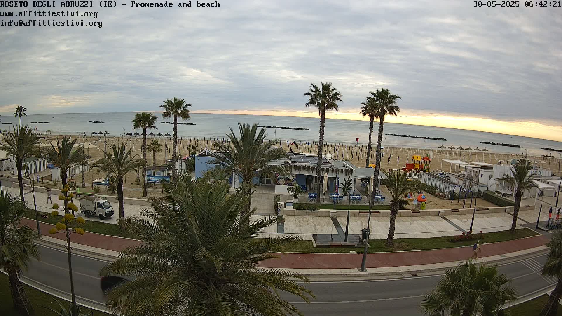 A seaside promenade and beach with palm trees, a small building, and a playground are visible under a cloudy sunrise.