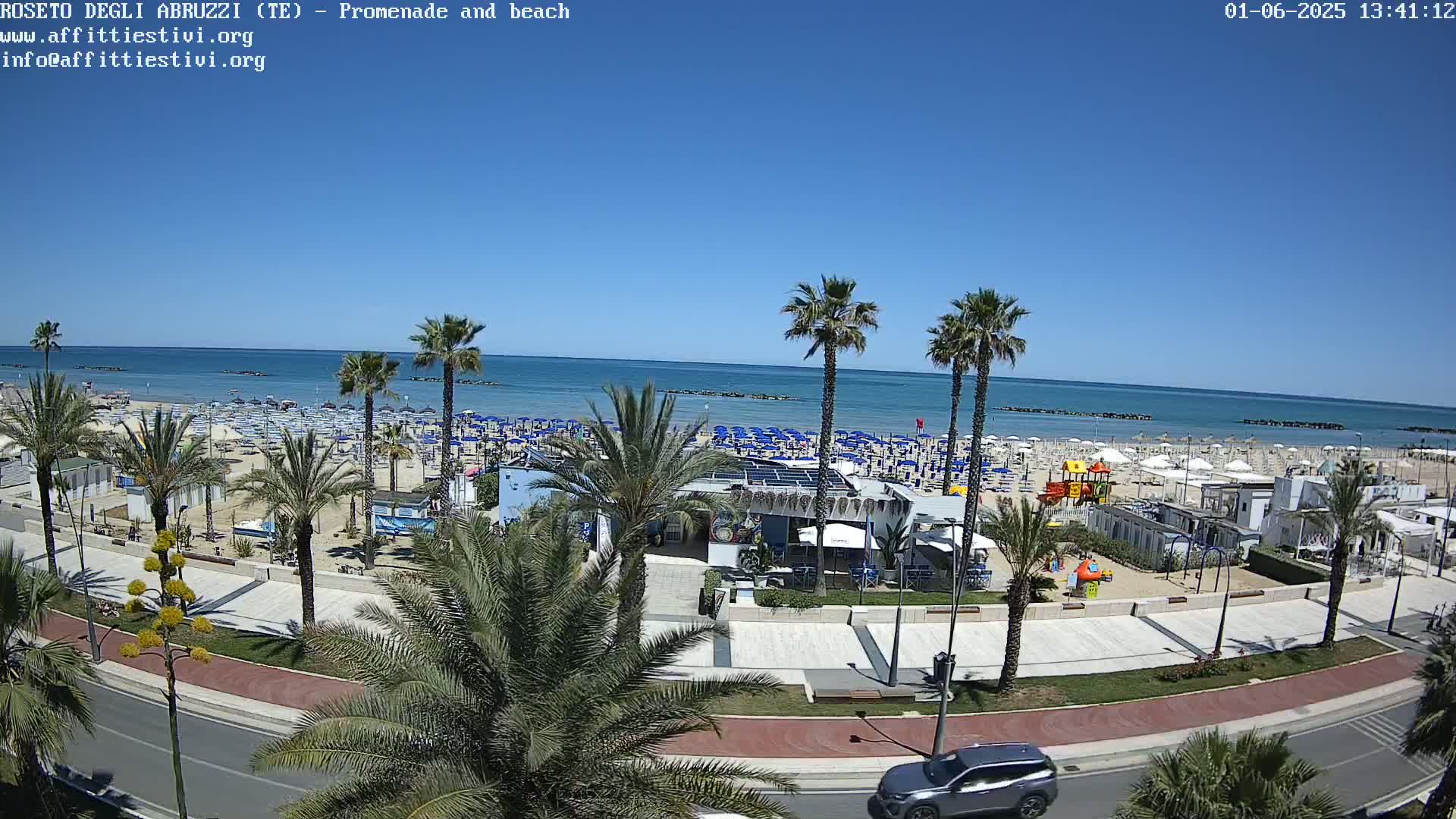 A sunny day at a beach, showing a promenade lined with palm trees, numerous beach umbrellas, and a calm ocean.