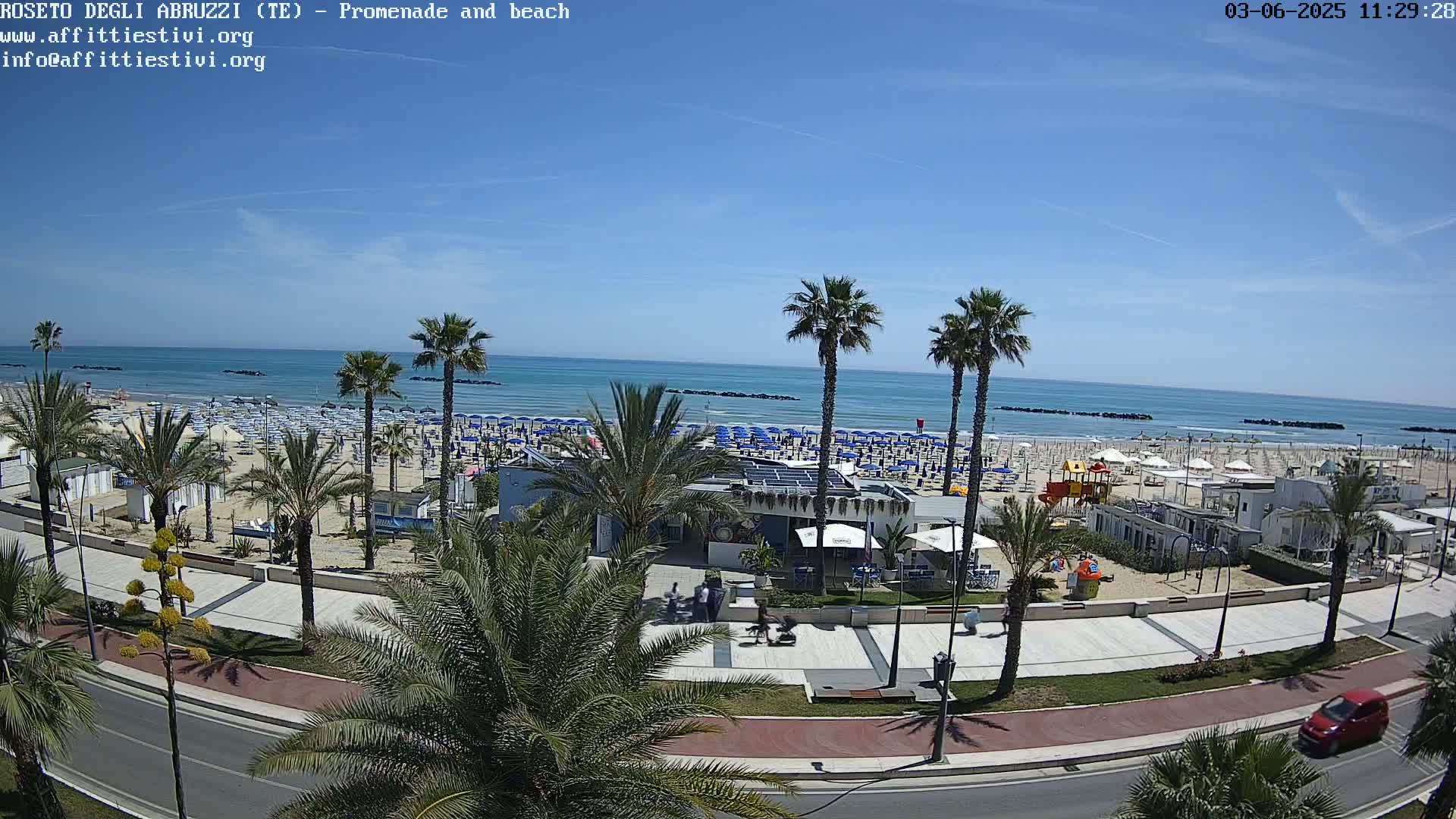 A sunny day at a beachside promenade shows a sandy beach lined with numerous blue umbrellas, palm trees, and beachfront buildings, all under a clear blue sky.