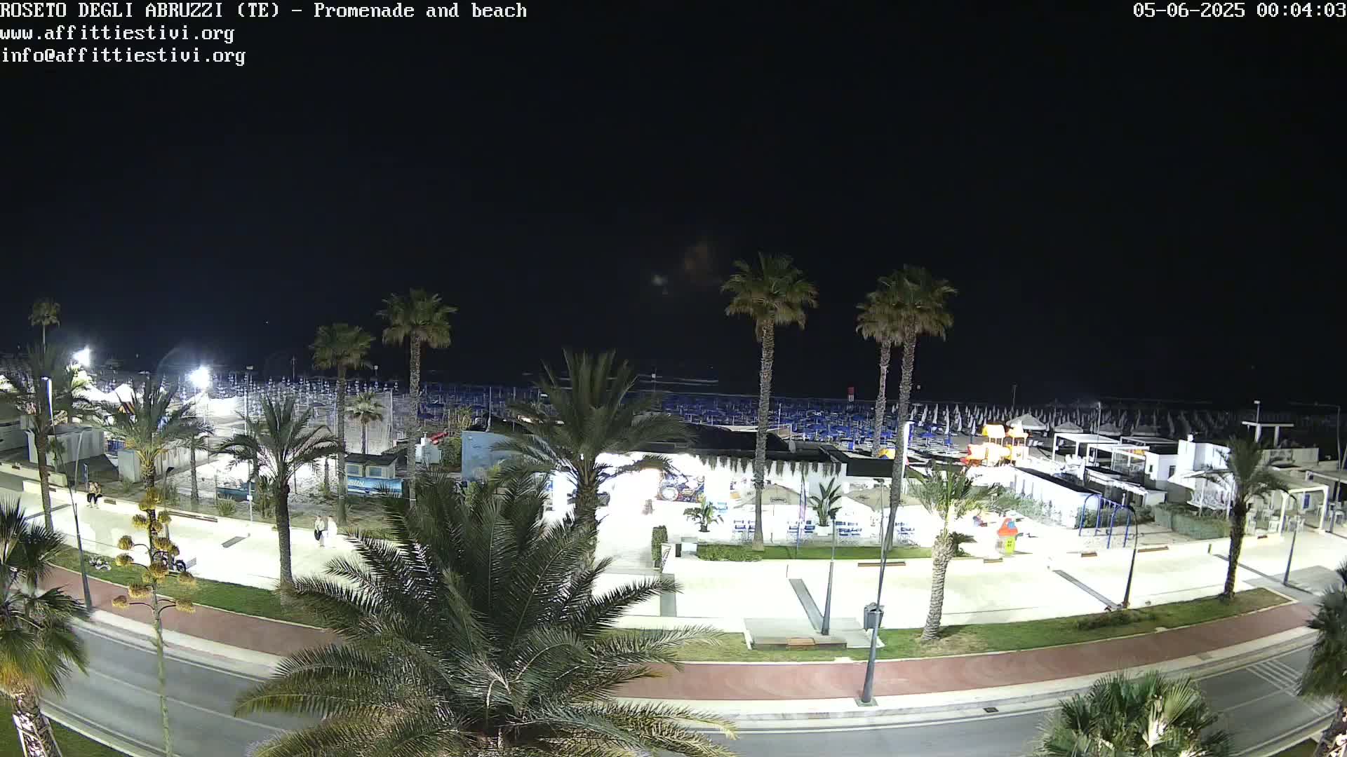 A nighttime, overhead view of a beach promenade and beach lined with palm trees, showing a mostly empty beach with some beach umbrellas and a few people on the boardwalk.