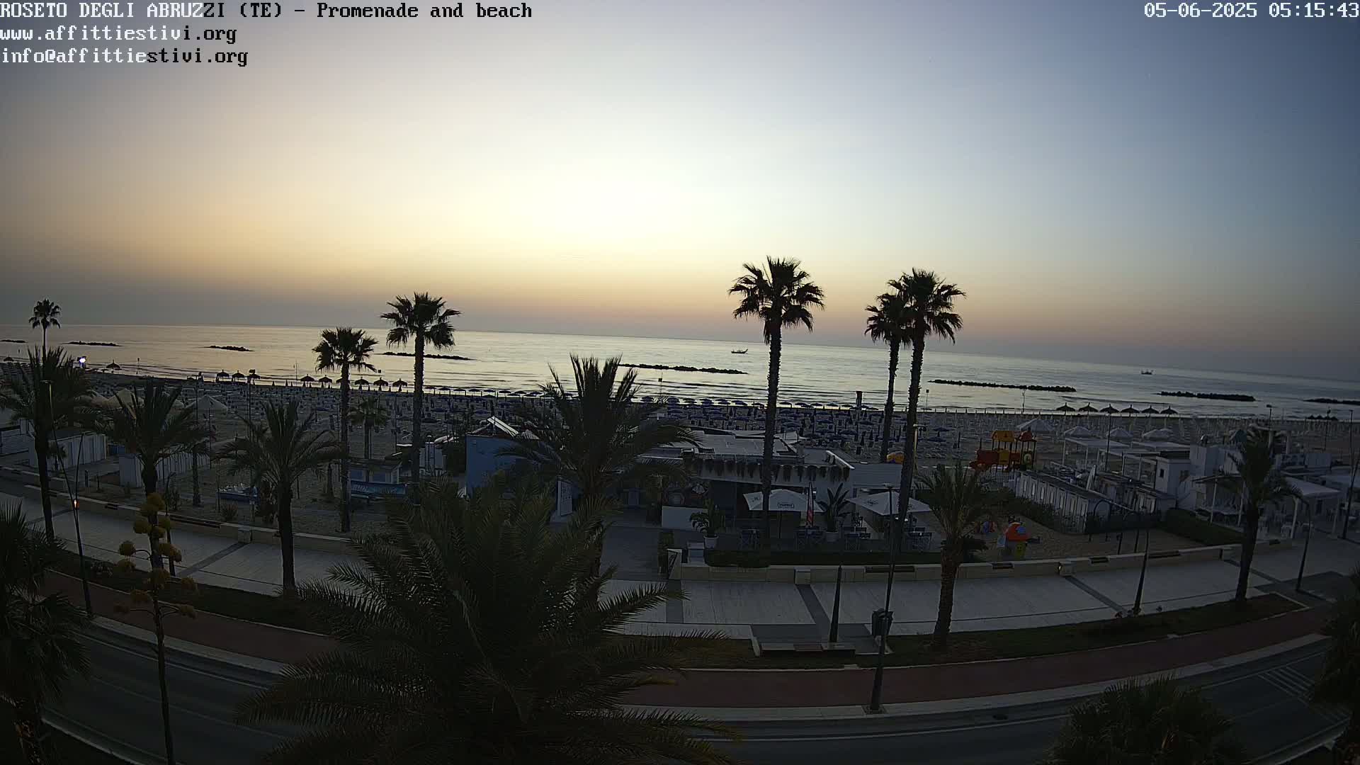 A beach with palm trees and beach umbrellas is visible at sunrise next to a paved promenade and road.