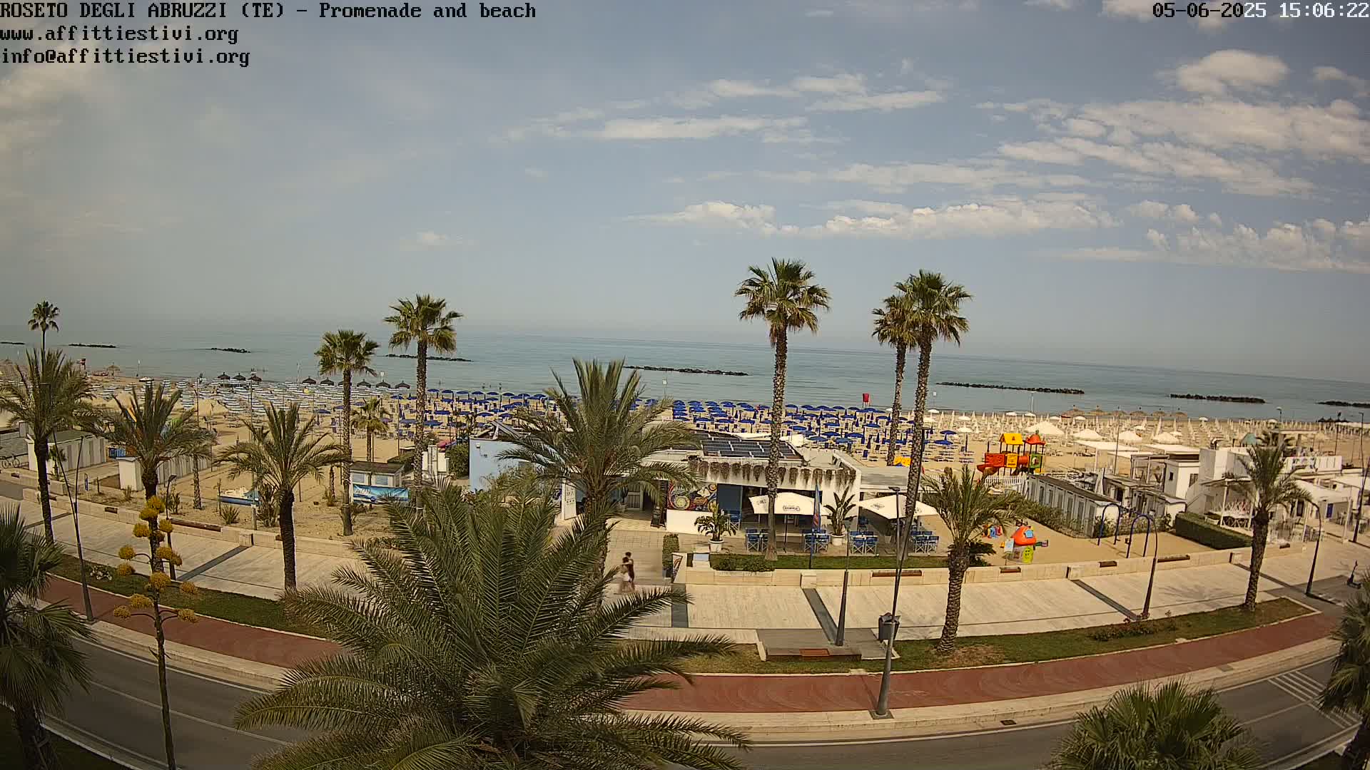 An aerial view shows a sunny beach with numerous blue umbrellas, palm trees lining a promenade beside a road, and a calm sea in the background.