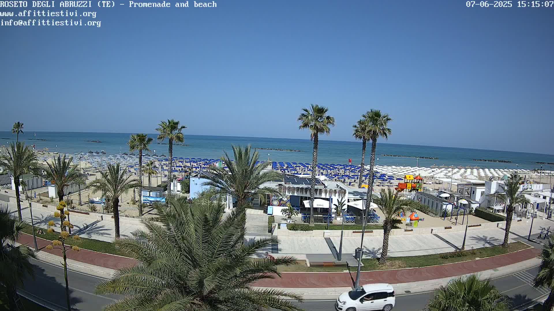 A sunny beach scene shows a promenade lined with palm trees, beach umbrellas, and a small beachfront restaurant, overlooking a calm ocean.