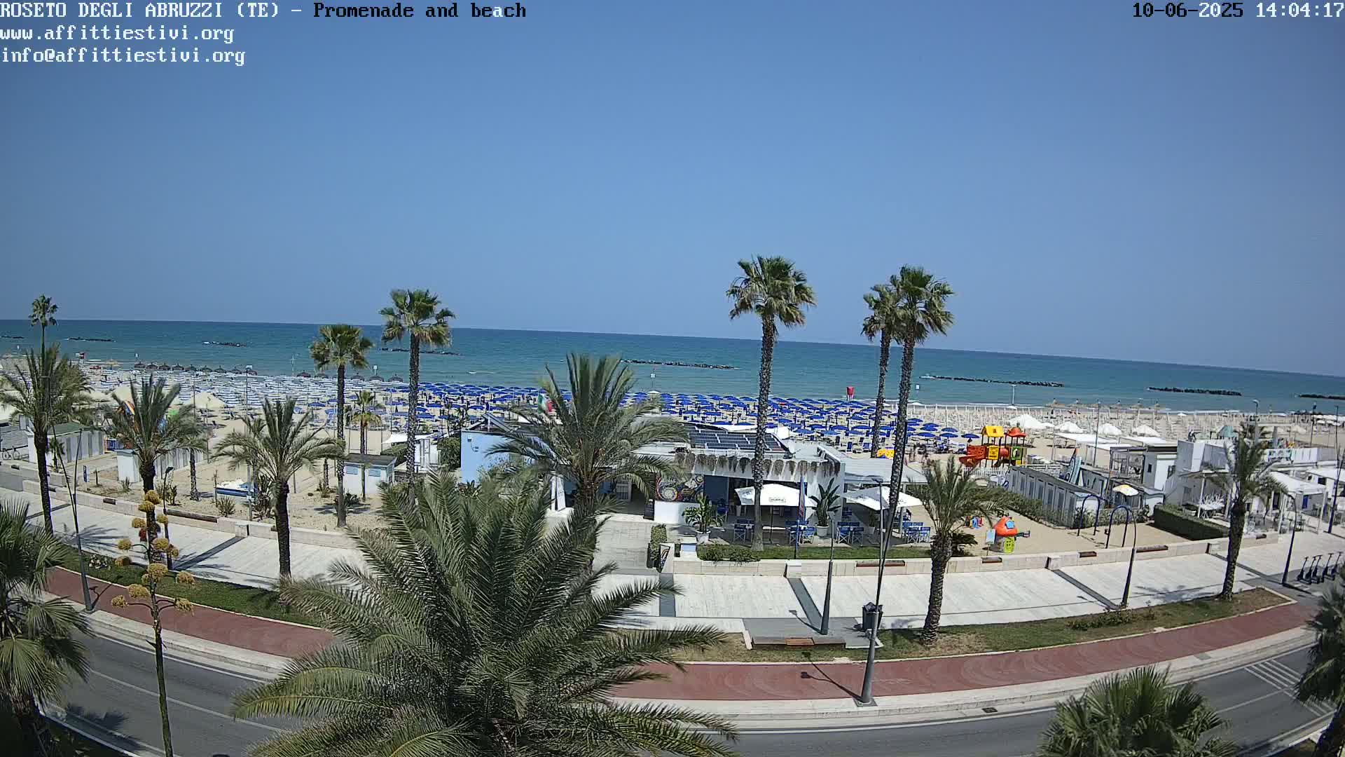 A beach with numerous blue umbrellas and a promenade lined with palm trees is visible under a clear, sunny sky.
