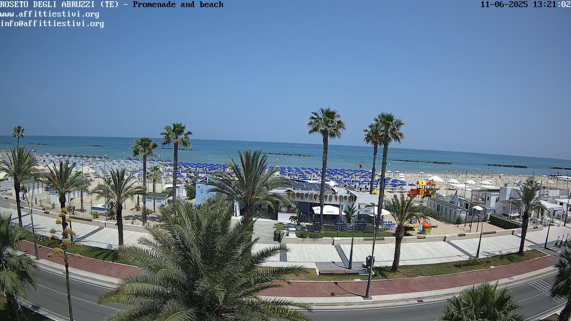 A sunny day at a beachside promenade shows numerous blue beach umbrellas lining a sandy beach, palm trees dotting the landscape, and a paved walkway separating the beach from the road.