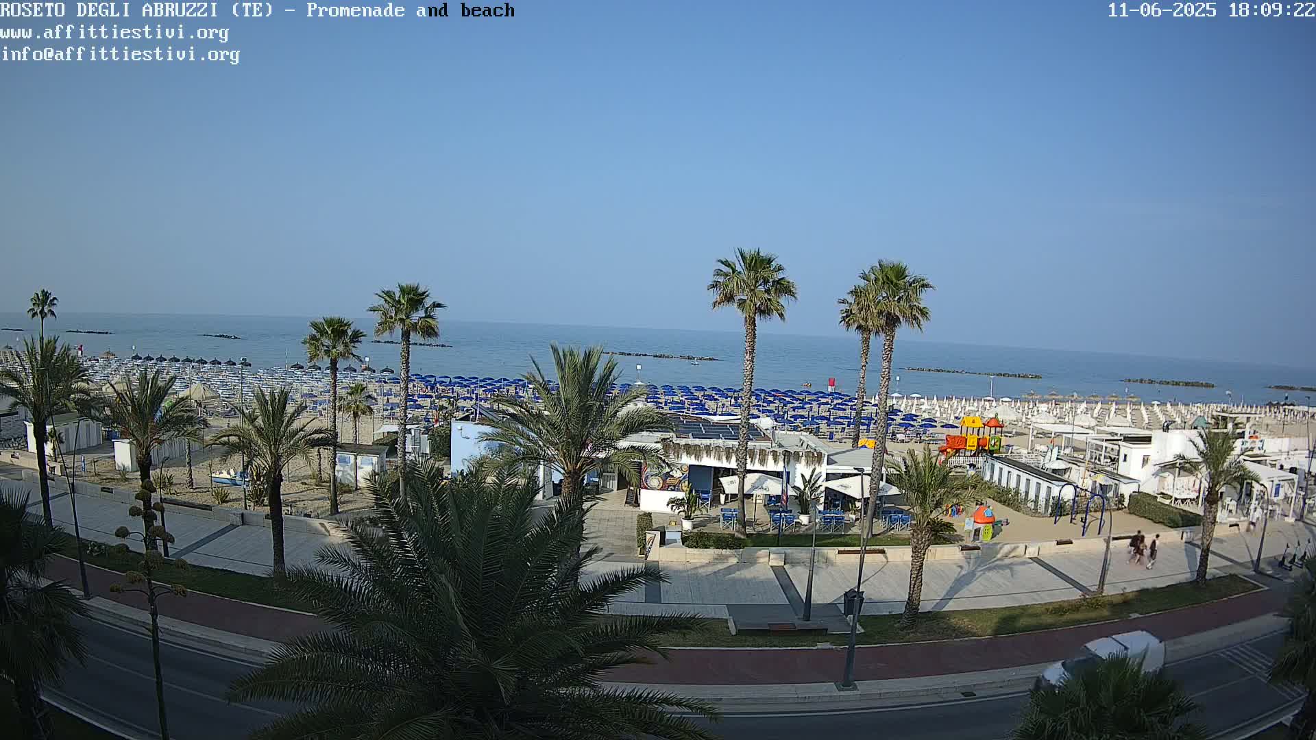 A sunny beach scene shows a wide sandy beach lined with rows of blue beach umbrellas, a promenade with palm trees, and a roadway with cars, all under a clear blue sky.