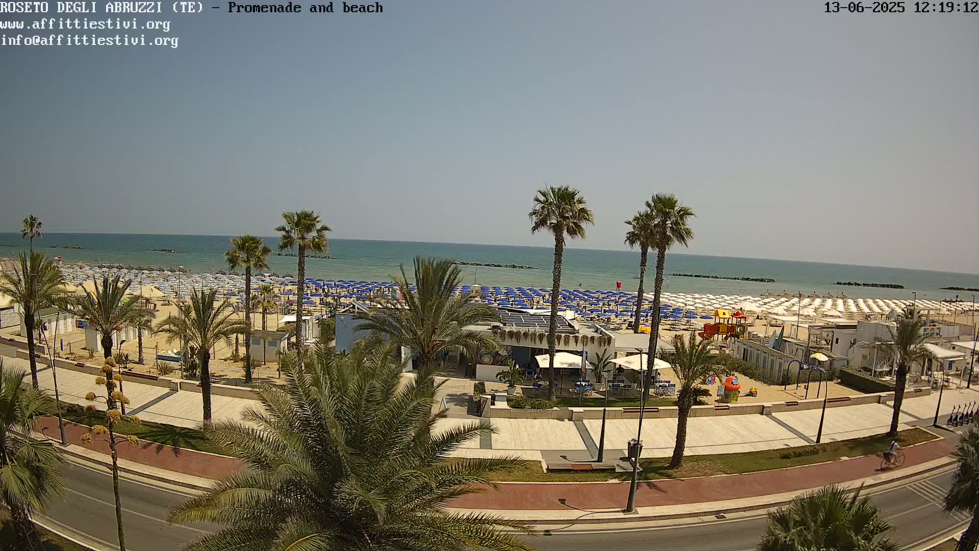 A sunny day at a beach, showing a crowded sandy shore lined with blue beach umbrellas, palm trees, and a paved promenade with a road next to it.