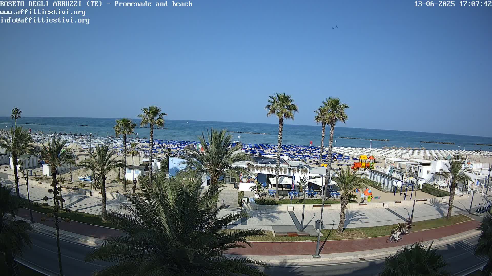 A beach with numerous blue and white umbrellas, palm trees, a promenade, and a beachfront restaurant is visible under a clear, sunny sky.