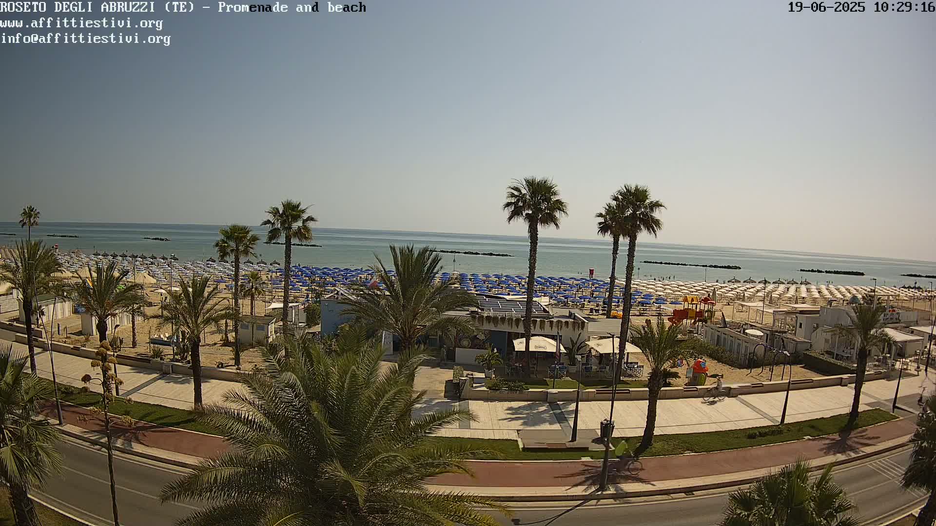 A beach with numerous blue umbrellas and palm trees lines a paved promenade next to a calm, light-blue sea under a clear sky.