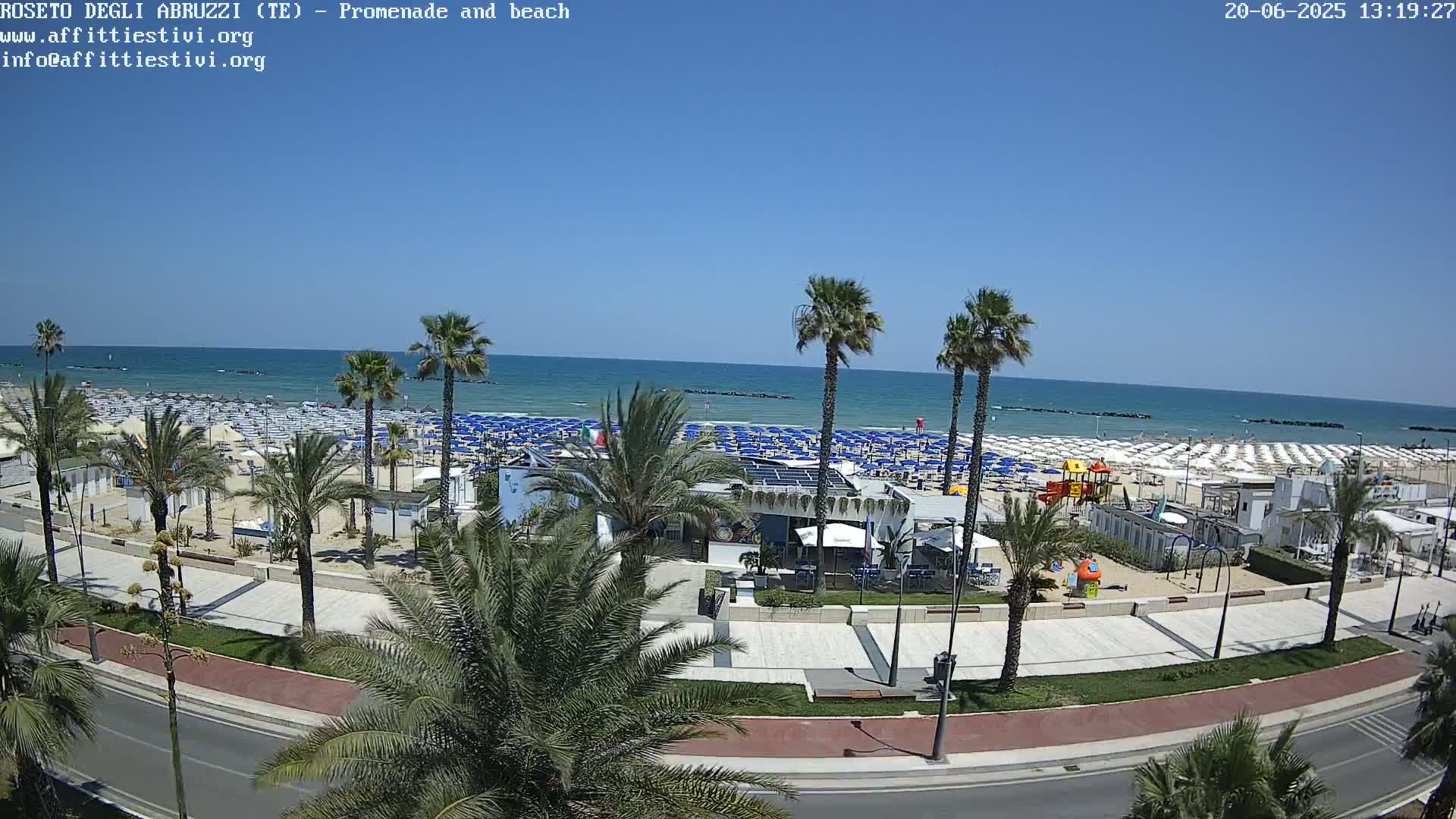 A sunny day at a beach with numerous umbrellas and palm trees lining a paved promenade beside a calm ocean.