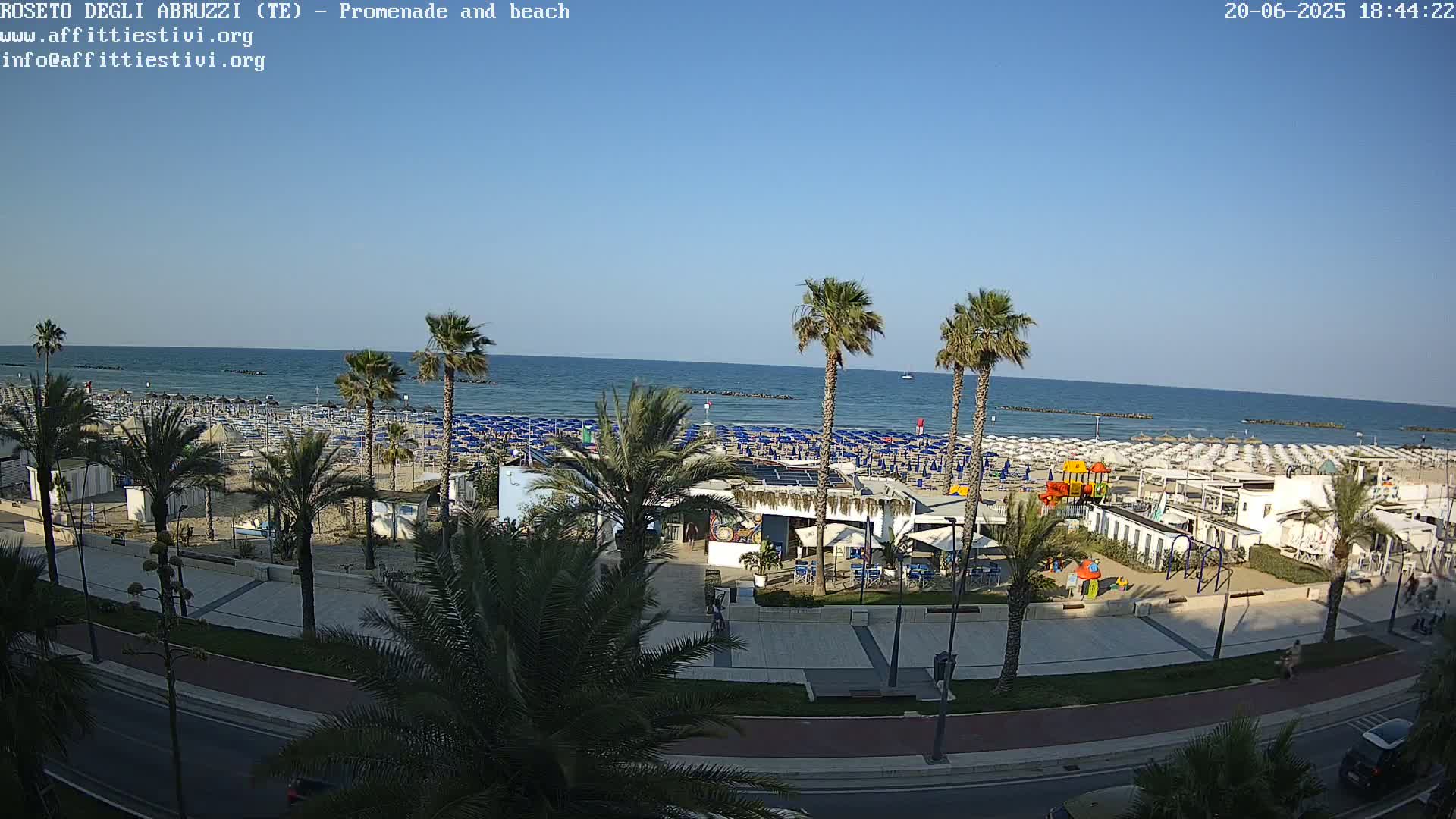 A sunny beach scene shows a long stretch of sand lined with rows of beach umbrellas, palm trees, a promenade, and a beachfront playground.