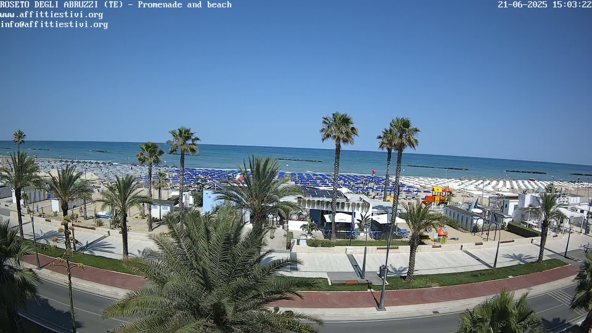 A sunny beach scene shows a promenade lined with palm trees, beach umbrellas, and small buildings, overlooking a calm ocean.