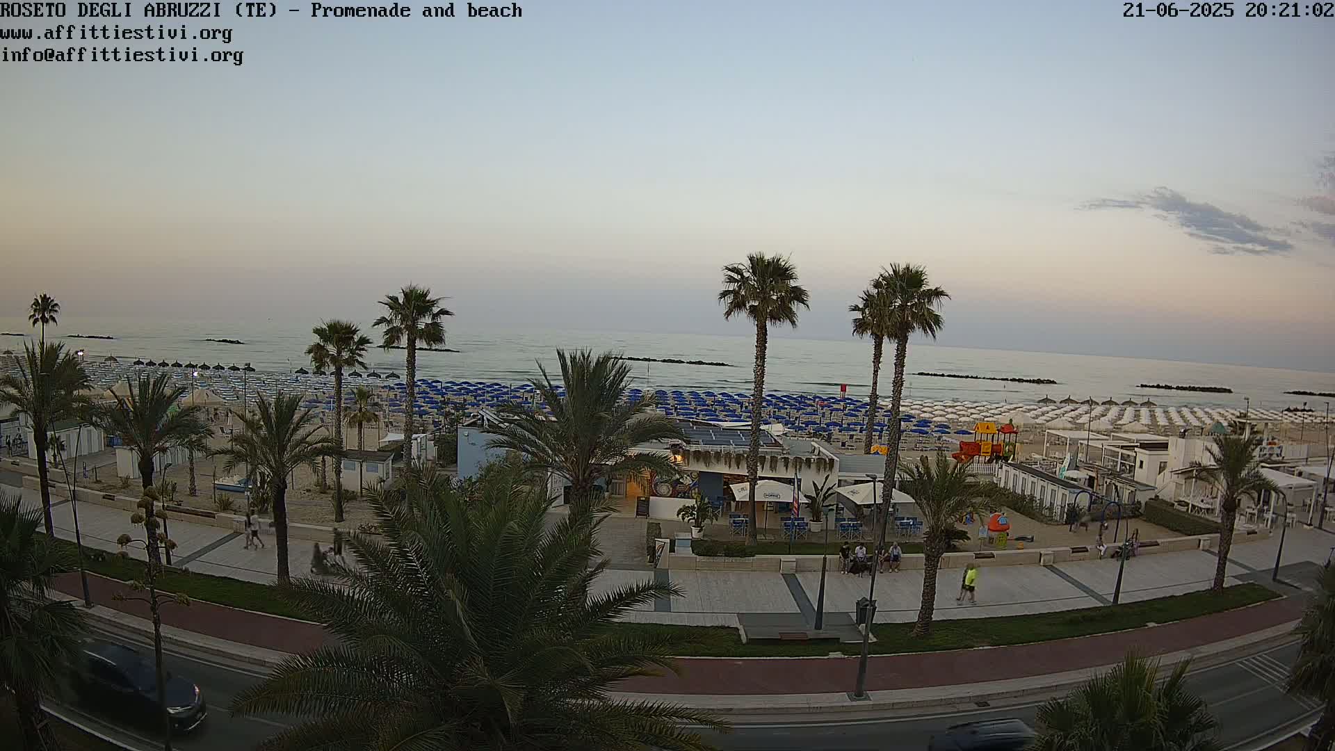 A beach with numerous blue and white umbrellas, palm trees, and a promenade at dusk under a mostly clear sky.