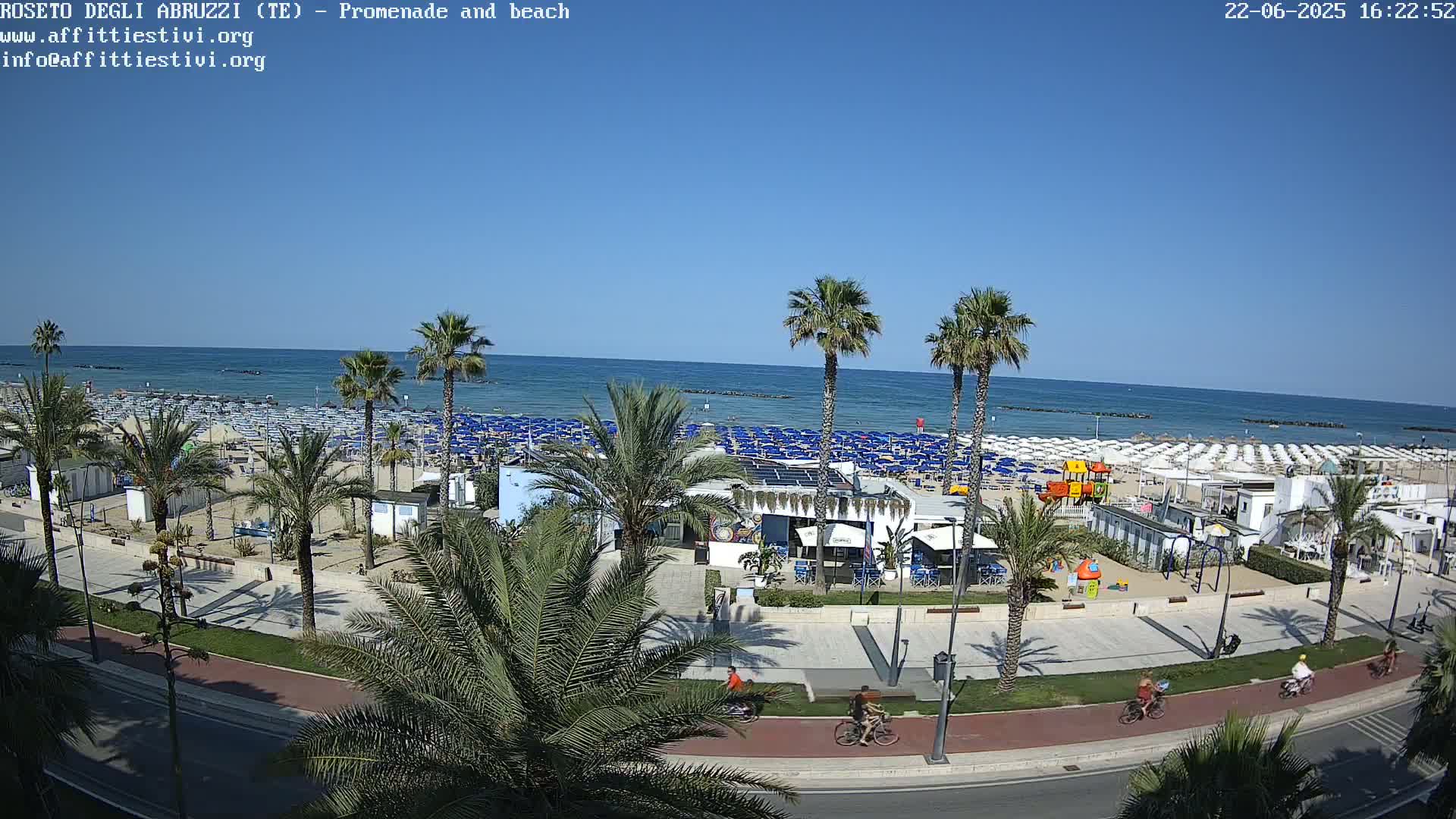 A beach with numerous blue beach umbrellas and palm trees is visible from a seaside promenade on a sunny day, with cyclists riding along a nearby path.